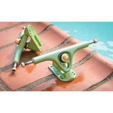 Two Paris V3 180mm 50-degree longboard trucks in Tropic Green with gold hardware on wet brick beside a pool. - Longboards USA